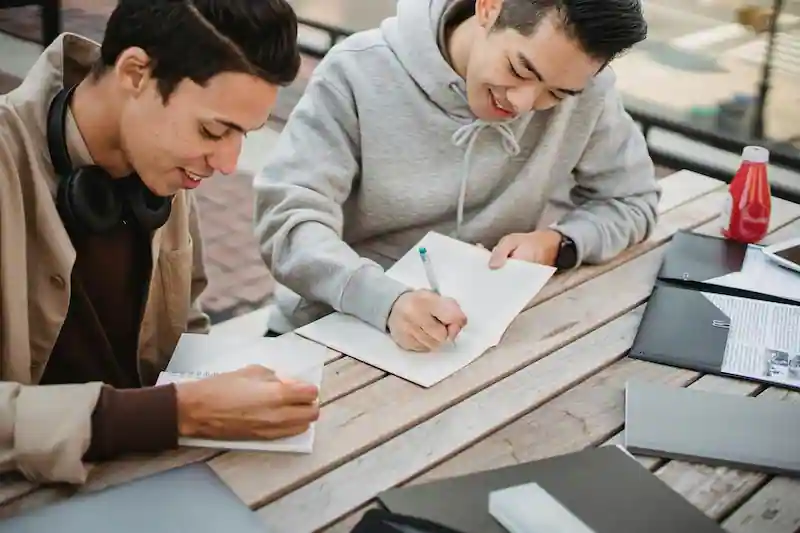 Dos jóvenes trabajando juntos al aire libre, tomando notas en sus libretas, simbolizando una gestión del tiempo efectiva y colaborativa para cumplir tareas y objetivos. mejorando en el bienestar emocional.